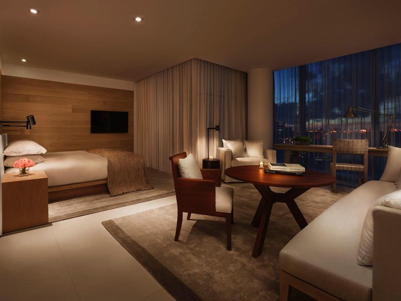 Modernes Hotelzimmer mit Wohnbereich, Bett, Fernseher und großer Fensterfront bei Abenddämmerung.