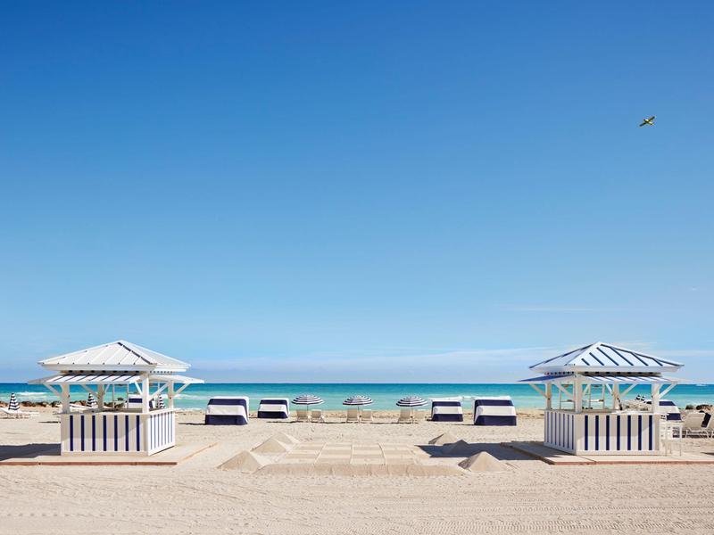 Sandstrand mit weißen Holzschirmen, blauer Himmel und ruhigem Meer im Hintergrund.