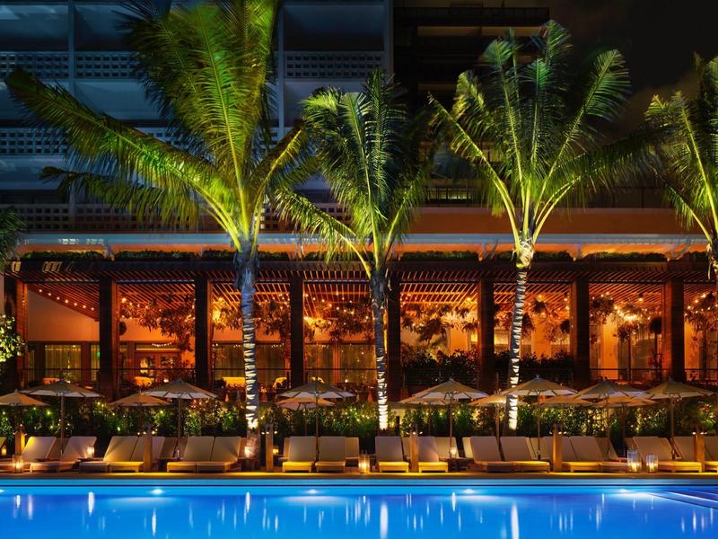 Abendliches Hotel mit beleuchteten Palmen und blauem Pool im Vordergrund.