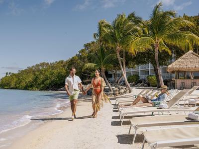 Paar läuft am sonnigen Strand mit Liegestühlen und Palmen entlang.