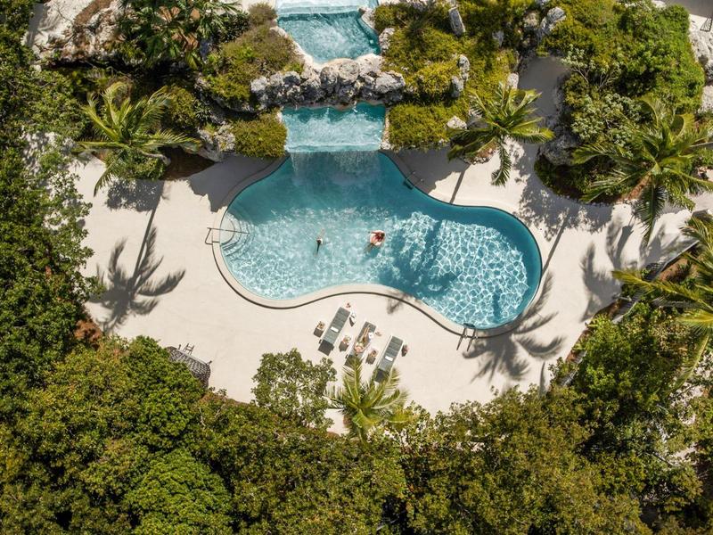 Schwimmbecken in organischer Form, umgeben von dichtem Grün und Palmen in tropischer Landschaft.