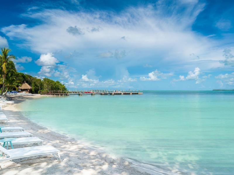 Weißer Sandstrand mit Liegestühlen, türkisfarbenem Wasser und blauem Himmel mit Wolken.