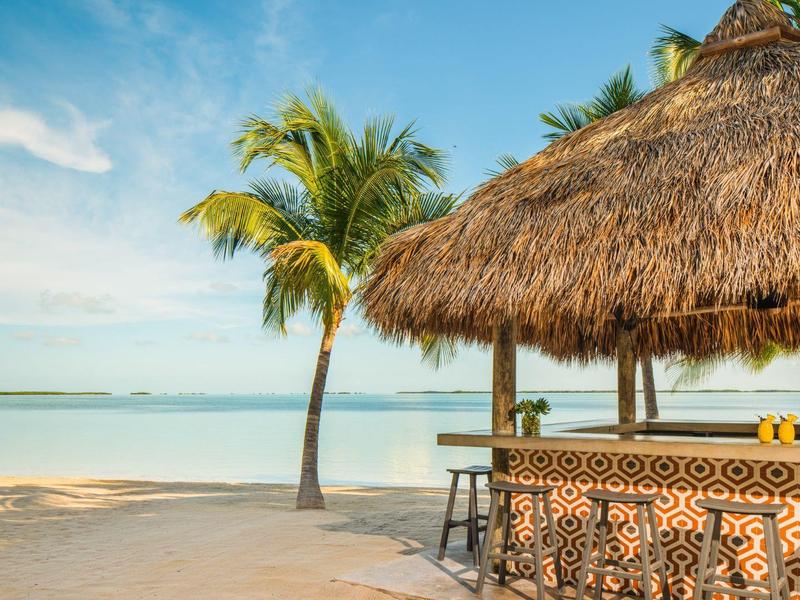 Strand mit Palmen, blauem Himmel und einer Bar mit Strohdach und gemusterten Fliesen.