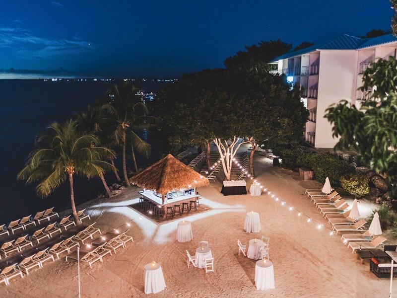 Strand mit weißen Strandstühlen, Palmen und beleuchtetem Pavillon bei Dämmerung.