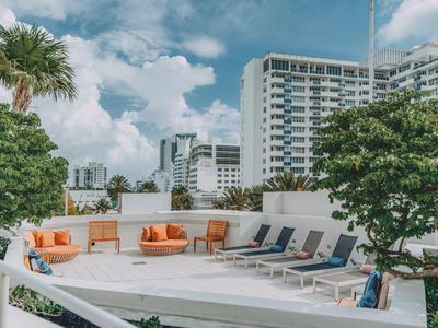 Moderne Hotelterrasse mit Sitzgelegenheiten, Liegestühlen und grünen Bäumen unter blauem Himmel.