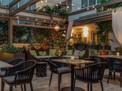 Gemütliche Hotelterrasse mit Stühlen, Tischen und grüner Pflanzendekoration unter Glasdach bei Abendlicht.