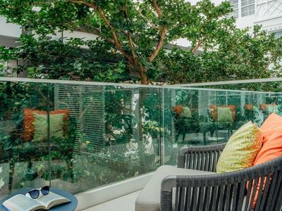 Balkon mit Stuhl, bunten Kissen und kleinem Tisch vor grünen Bäumen in Hotelumgebung.