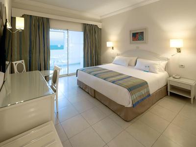 Hell eingerichtetes Hotelzimmer mit Doppelbett, Schreibtisch und Blick auf das Meer.