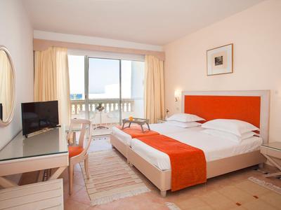 Jasny pokój hotelowy z podwójnym łóżkiem, pomarańczowymi akcentami i balkonem z widokiem na morze.