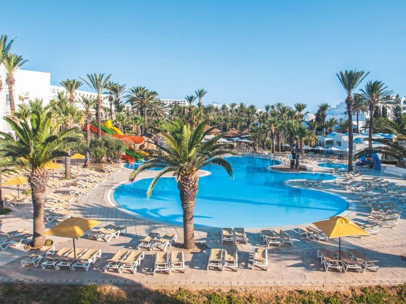 Großer Pool mit Palmen und Liegestühlen unter blauem Himmel an einem Hotel.