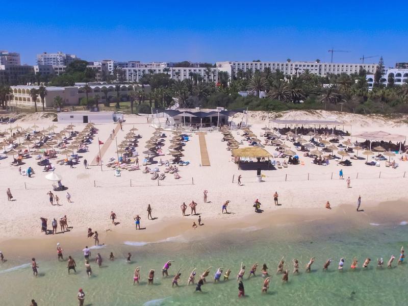 Breiter Sandstrand mit Liegestühlen und vielen Menschen am klaren Meer unter blauem Himmel.