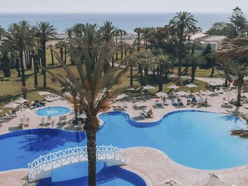 Großer Außenpool mit Sonnenliegen, Palmen und Blick auf das Meer in einem Hotelresort.