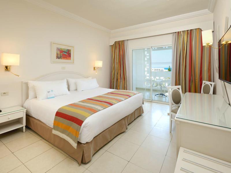 Helles Hotelzimmer mit Doppelbett, Fernseher und Balkon mit Meerblick.