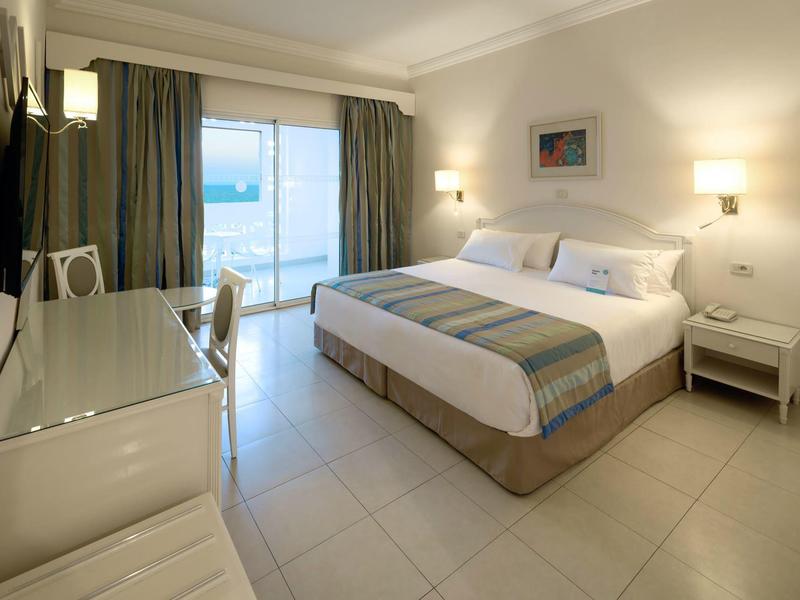 Hell eingerichtetes Hotelzimmer mit Doppelbett, Schreibtisch und Blick auf das Meer.