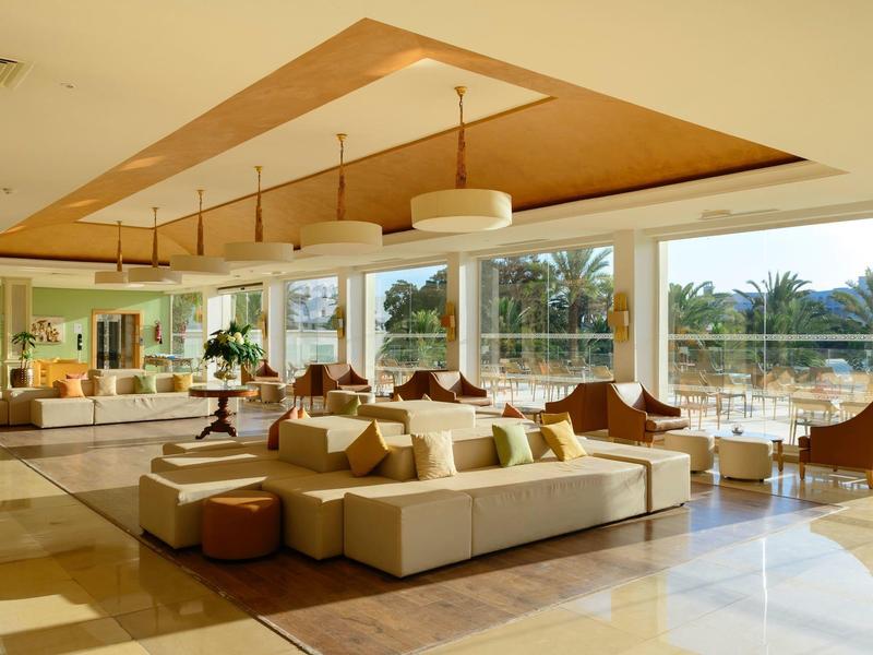 Helles Hotel-Lounge mit großen Fenstern, moderner Sitzgruppe und Ausblick auf Palmen.