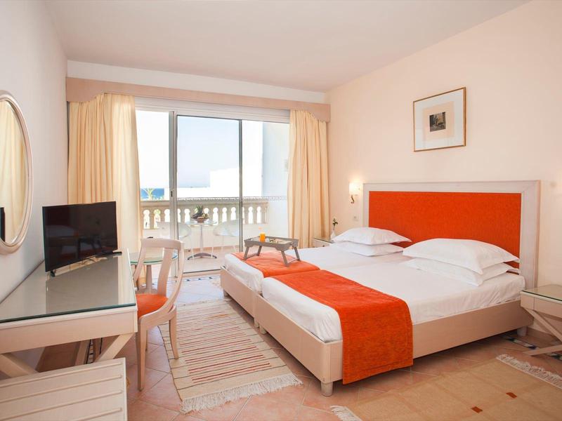 Helles Hotelzimmer mit Doppelbett, orangem Bettläufer, TV und Balkon mit Meerblick.