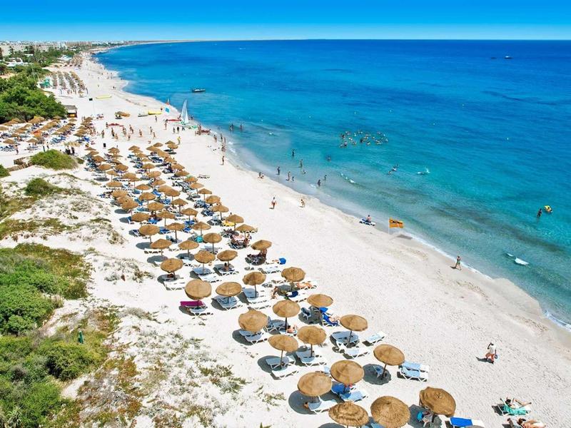 Weißer Sandstrand mit blauen Sonnenschirmen, türkisblaues Meer und Spaziergänger am Ufer.