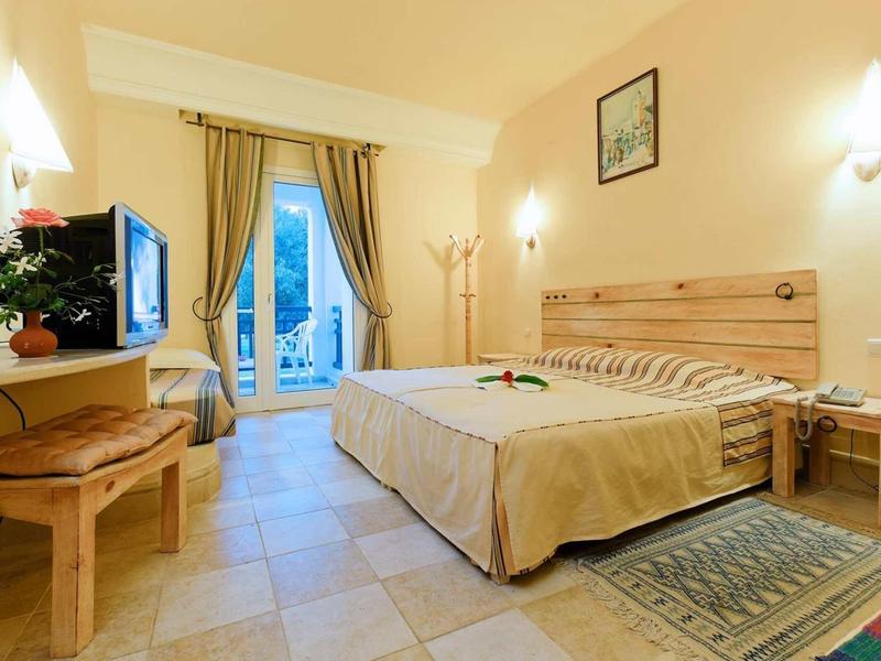 Helles Hotelzimmer mit Doppelbett, Fernseher, Gardinen und Zugang zu einem Balkon bei Tageslicht.