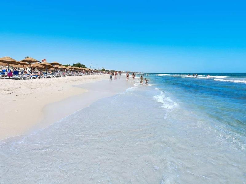 Breiter Sandstrand mit Sonnenschirmen, Menschen und klarem, blauem Meer unter blauem Himmel.