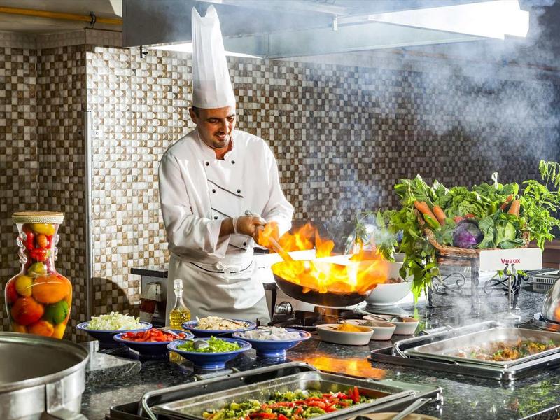 Koch mit weißer Mütze flambiert Speisen in einer professionellen Küchenumgebung mit vielen frischen Zutaten.