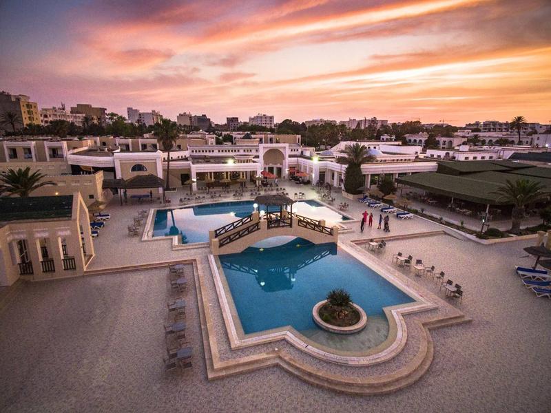 Großes, gemütliches Hotel mit Pool, Sonnenliegen und farbenfrohem Himmel bei Sonnenuntergang.