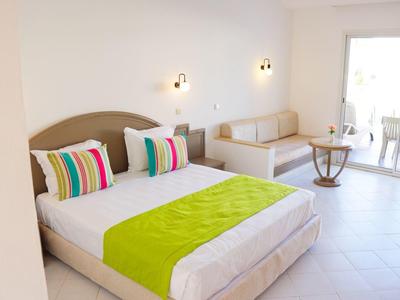 Helles Hotelzimmer mit Doppelbett, bunten Kissen und Balkon mit Sitzecke.