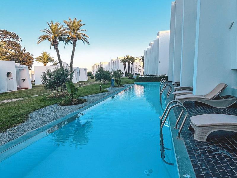 Long pool with white buildings and palm trees under clear blue sky.