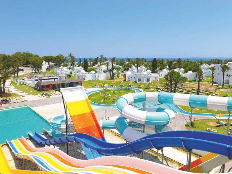 Colorful water slides and a pool in a sunny resort with white buildings and palm trees in the background.