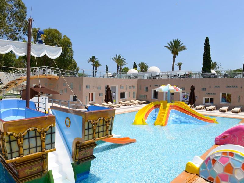 Outdoor pool with colorful slides and pirate ship play structure under clear blue sky.