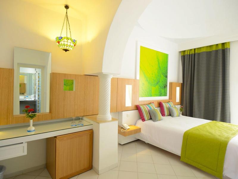 Bright hotel room with white bed, green accents, a vanity desk, and modern lighting.