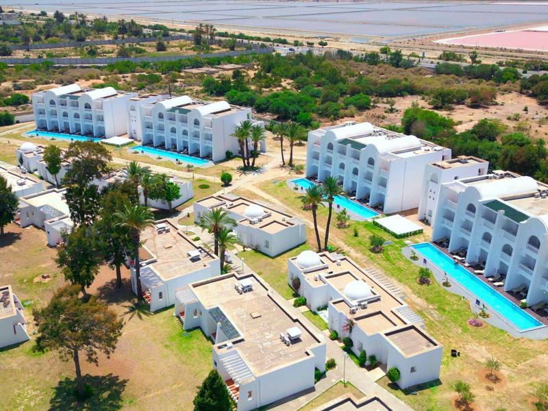 Aerial view of a resort with white buildings, pool, and green trees in a semi-arid landscape.