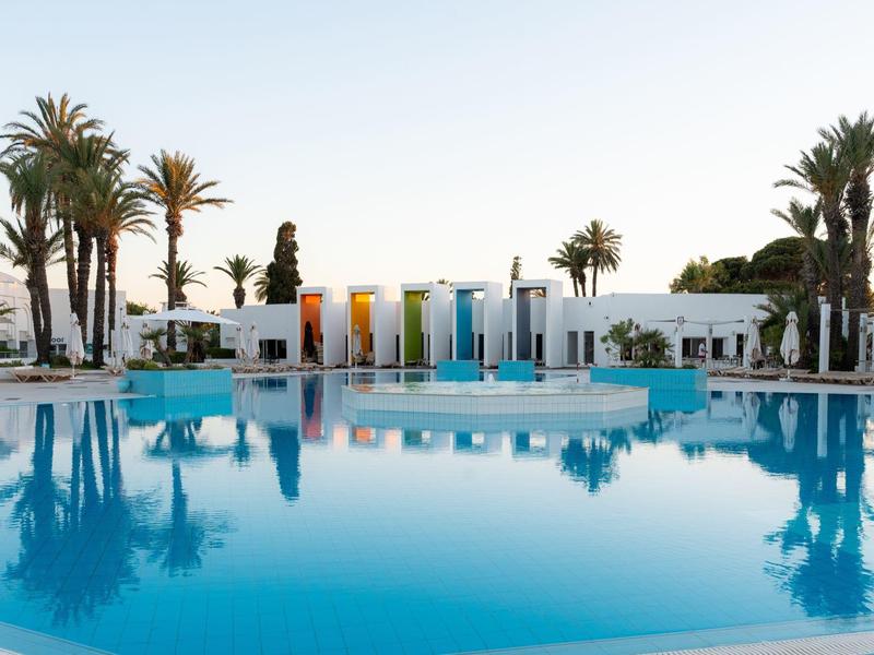 Large hotel pool with colorful umbrellas and palm trees around under clear sky.