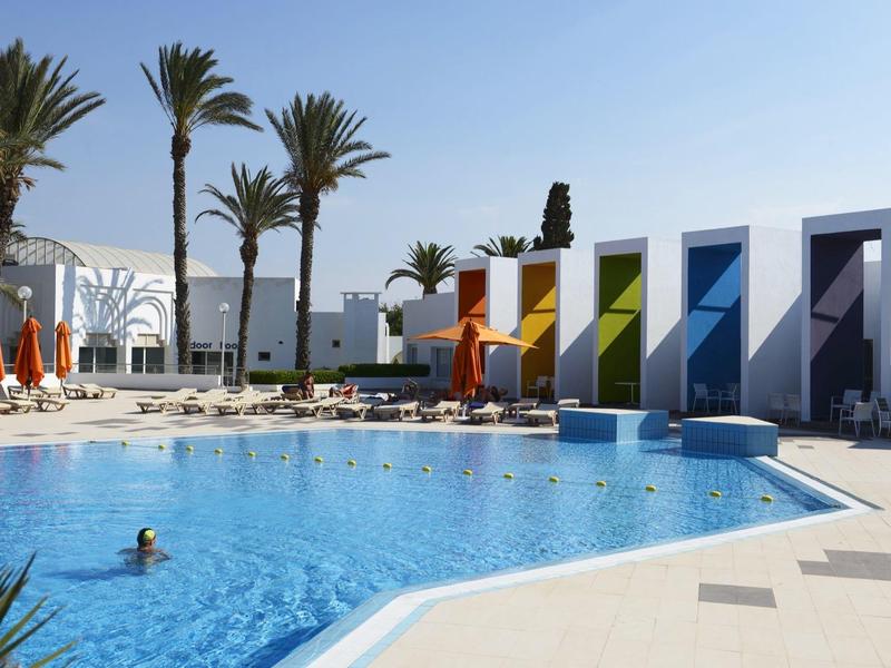 Modern hotel pool with colorful cabins, palm trees, and umbrellas under clear sky.
