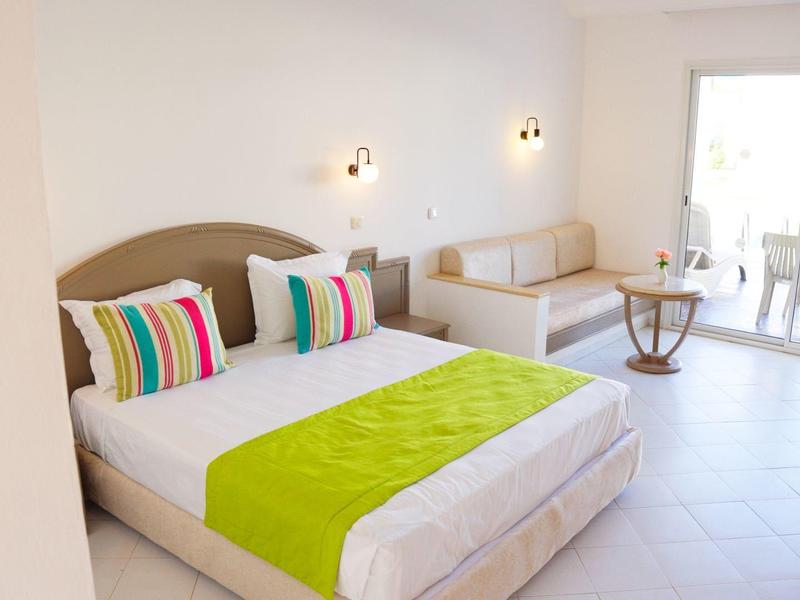 Bright hotel room with double bed, colorful pillows, and balcony seating area.