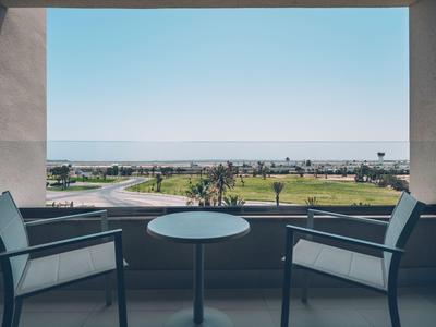 Balkon mit zwei Stühlen und rundem Tisch, Blick auf Küstenlandschaft und blauen Himmel.