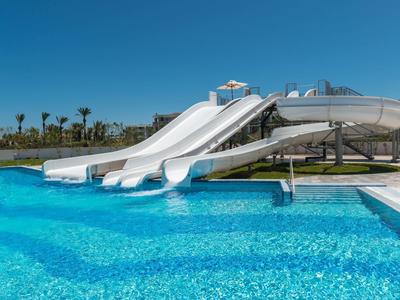 Drei weiße Wasserrutschen führen in ein klares, blaues Schwimmbecken unter blauem Himmel.