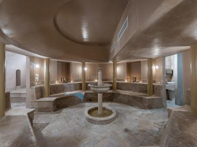 Großer, runder Hamam mit zentralem Brunnen, warmen Farben und Sitzbänken aus Stein.