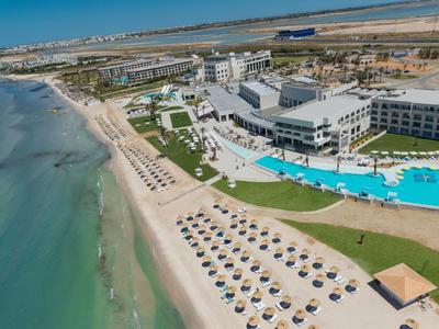 Hotelanlage mit großen Pools, Sonnenliegen am Sandstrand und Küstenlinie unter blauem Himmel.