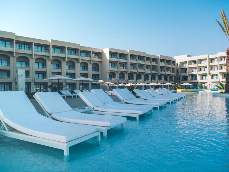 Hôtel avec piscine et chaises longues blanches sous un ciel clair.