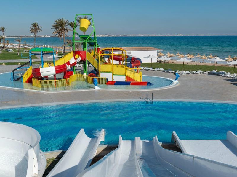 Parc aquatique avec toboggans colorés, piscine et mer en arrière-plan par jour ensoleillé.