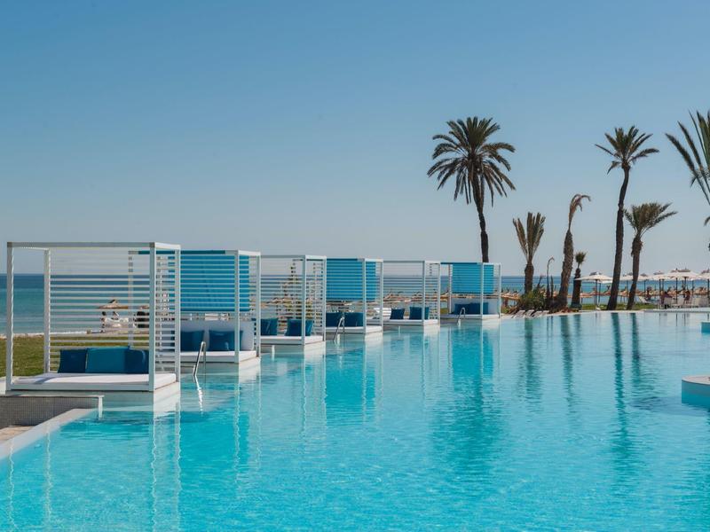 Piscine moderne avec auvents blancs et palmiers au bord de la mer sous un ciel clair.