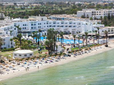 Complejo hotelero con edificios blancos, piscinas y playa de arena junto a un mar tranquilo.