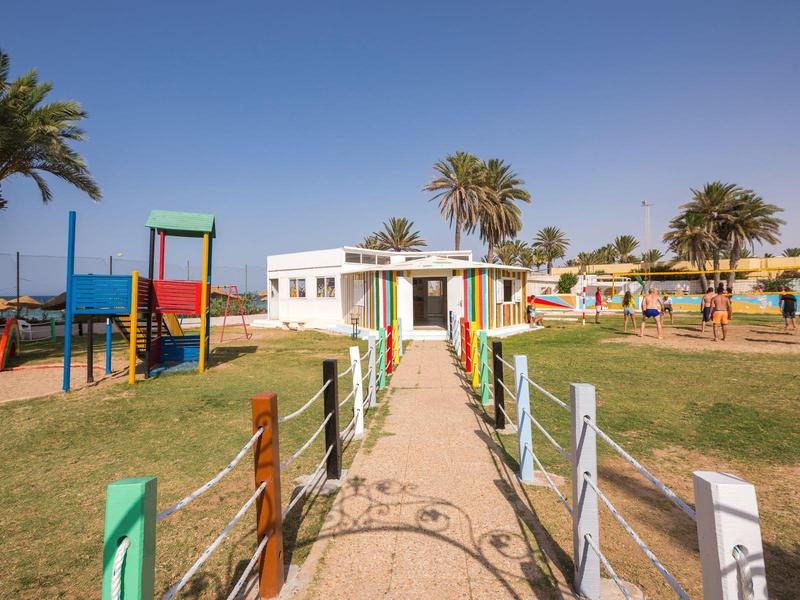 Camino con valla colorida que conduce a un parque infantil con toboganes y palmeras al fondo.
