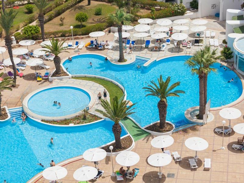 Área de piscina con mesas blancas redondas, sombrillas y palmeras junto a una gran piscina del hotel.