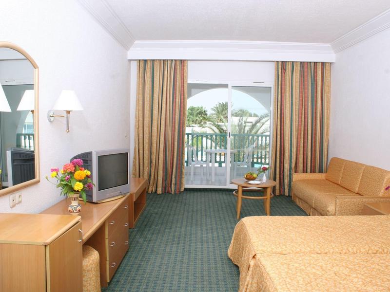 Habitación de hotel luminosa con cama doble, TV, sofá y balcón con vistas a palmeras.