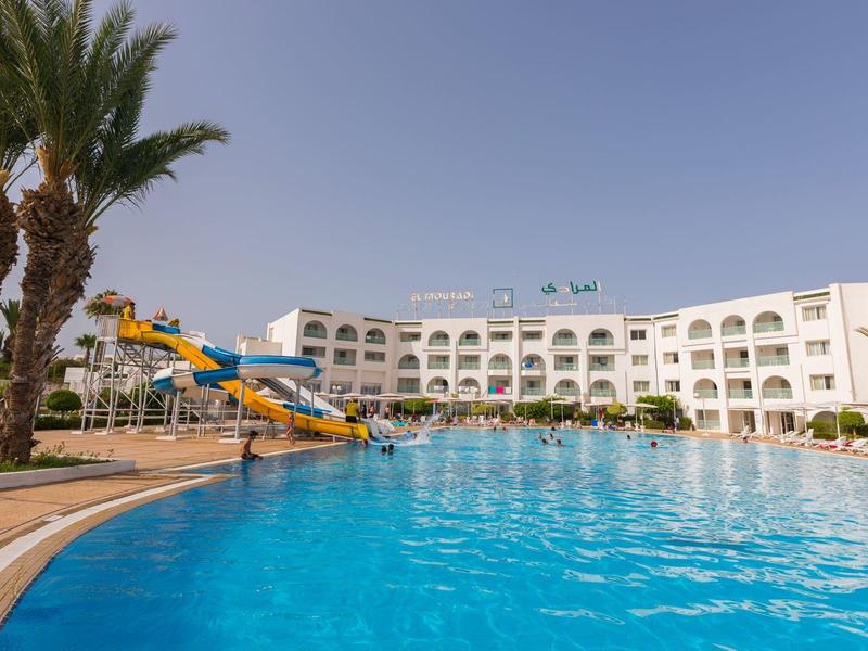 Gran piscina al aire libre frente a un hotel de varios pisos con tobogán de agua y palmeras.