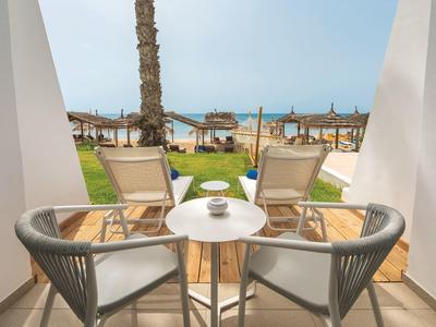 Blick durch weißen Balkon auf Stühle, Tisch, Rasen, Palmen und Meer im Hintergrund bei blauem Himmel.