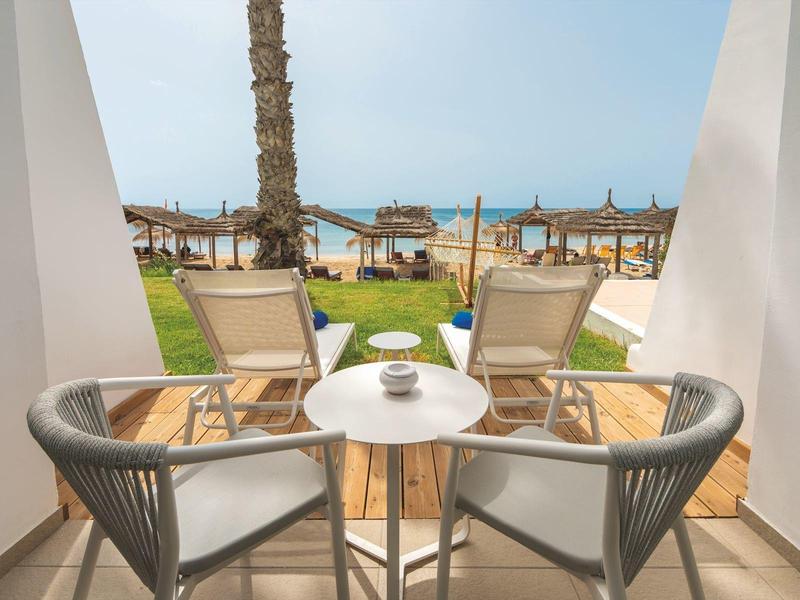 Blick durch weißen Balkon auf Stühle, Tisch, Rasen, Palmen und Meer im Hintergrund bei blauem Himmel.