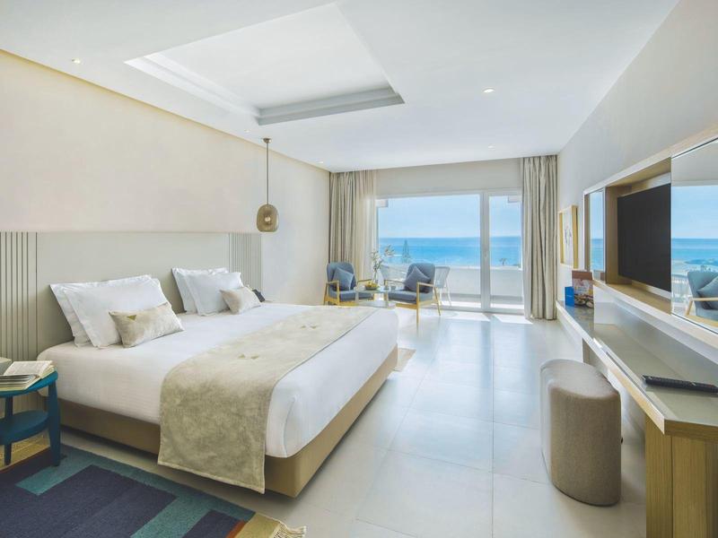 Modernes Schlafzimmer mit großem Bett, Meerblick durch Glasfenster und heller, minimalistischer Einrichtung.