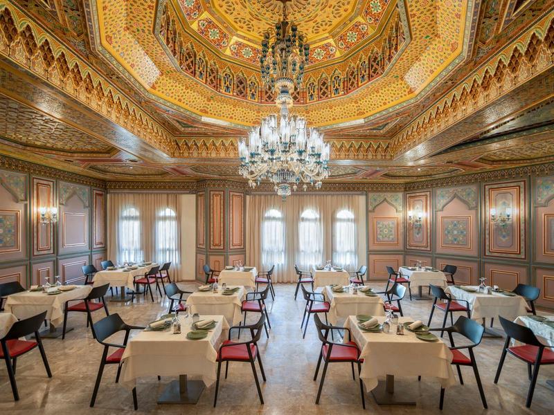 Großer prunkvoller Saal mit goldener Decke, Kronleuchtern, Tischen mit weißen Tüchern und roten Stühlen.